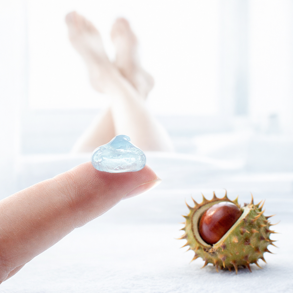Ein kühles, leicht bläuliches oder transparentes Gel auf einer Fingerspitze, im Hintergrund die Silhouette von hochgelegten Beinen.